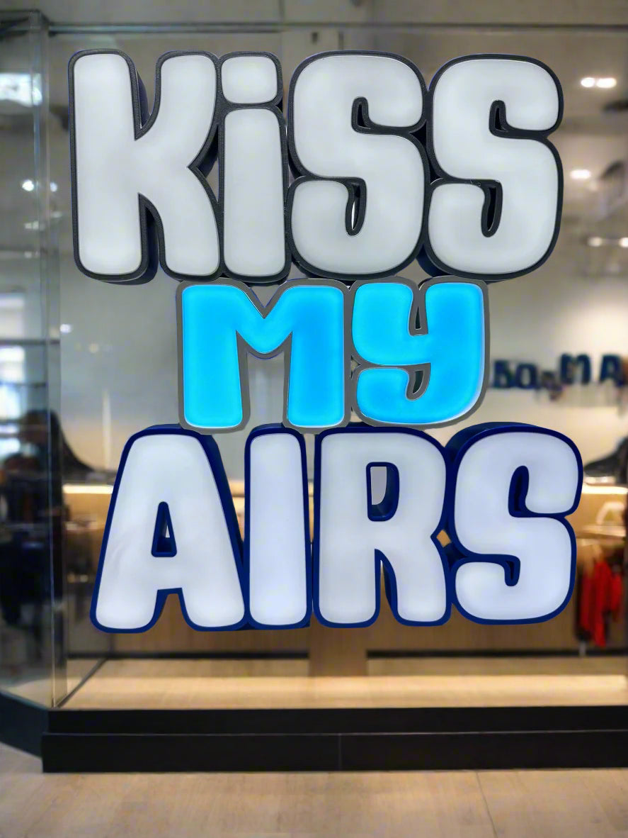 Front view of the Kiss My Airs LED Channel Letter Sign, featuring bold white letters with black outlines and a light blue 'MY' in the center, illuminated against a white background, perfect for home or office decor.