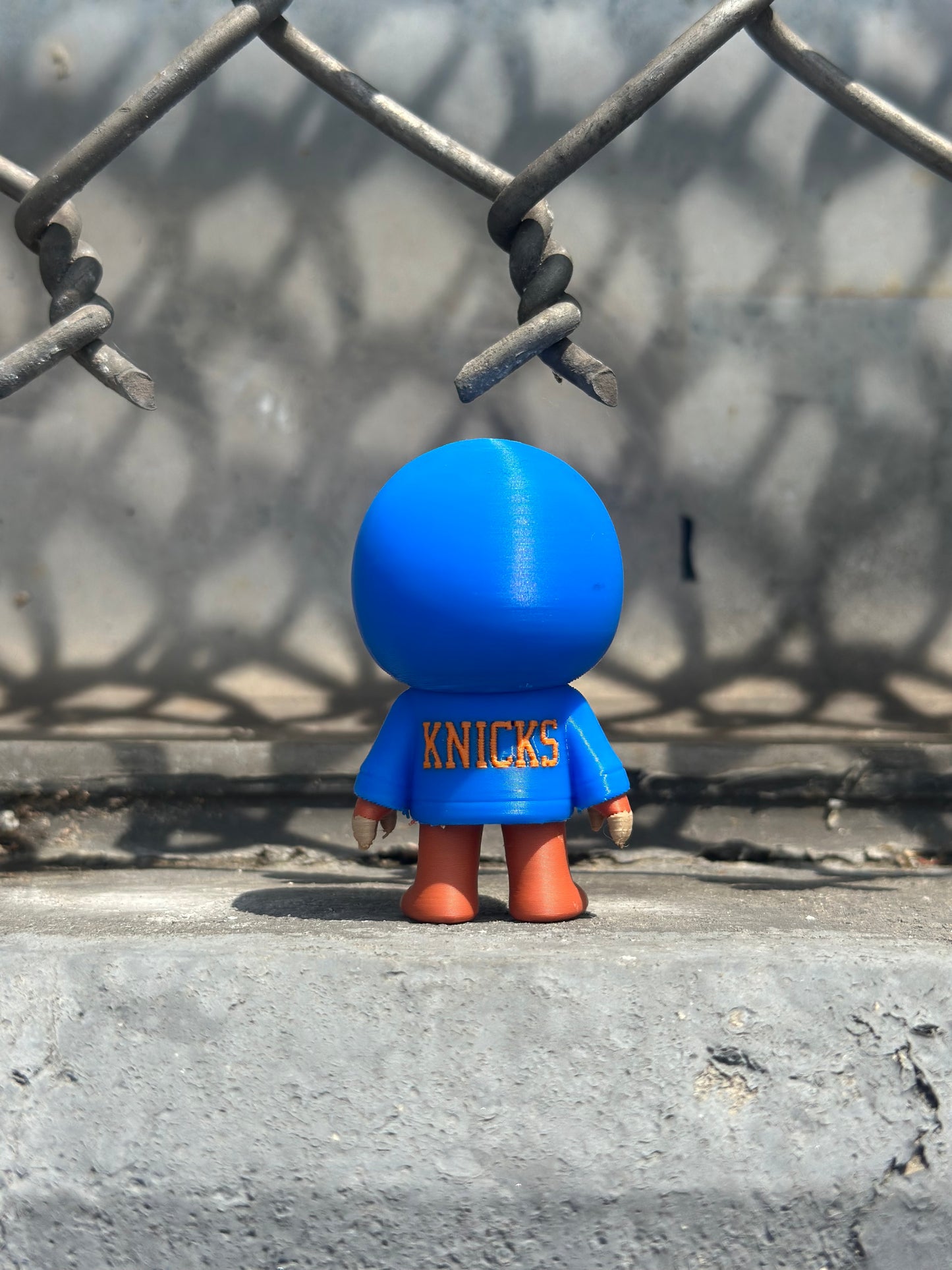 Blue toy figure with 'Knicks' branding peeking through a chain-link fence.