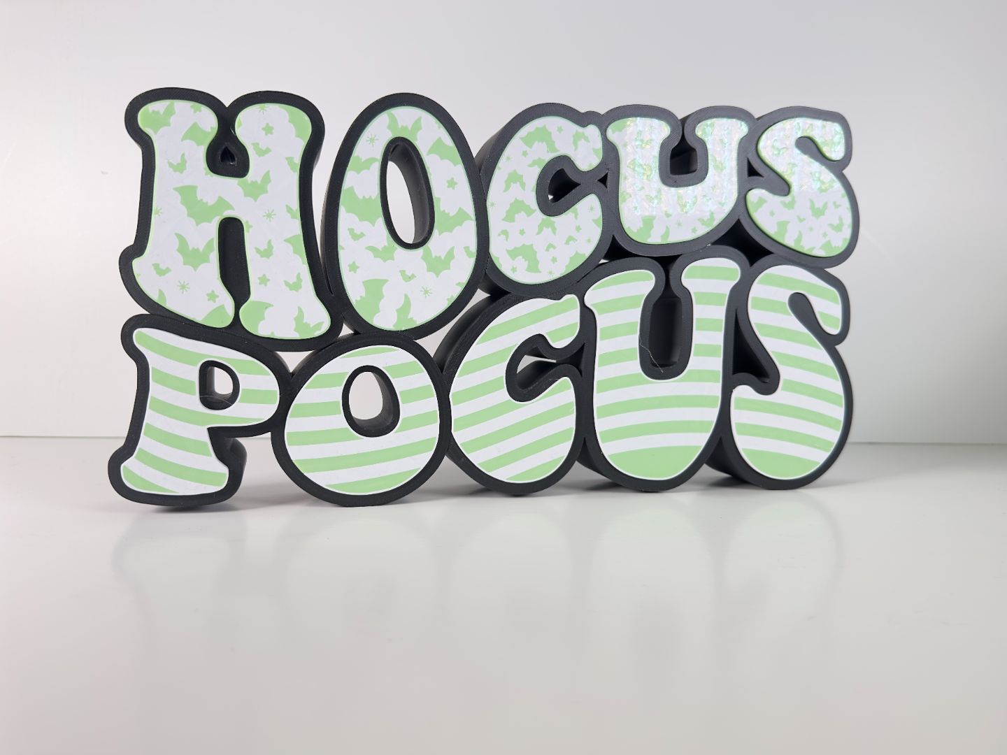 A “Hocus Pocus” LED sign with green bat patterns and striped letters, resting on a white background, perfect for Halloween decor