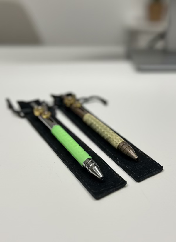 Two pens with decorative elements on a gray surface with tools in the background