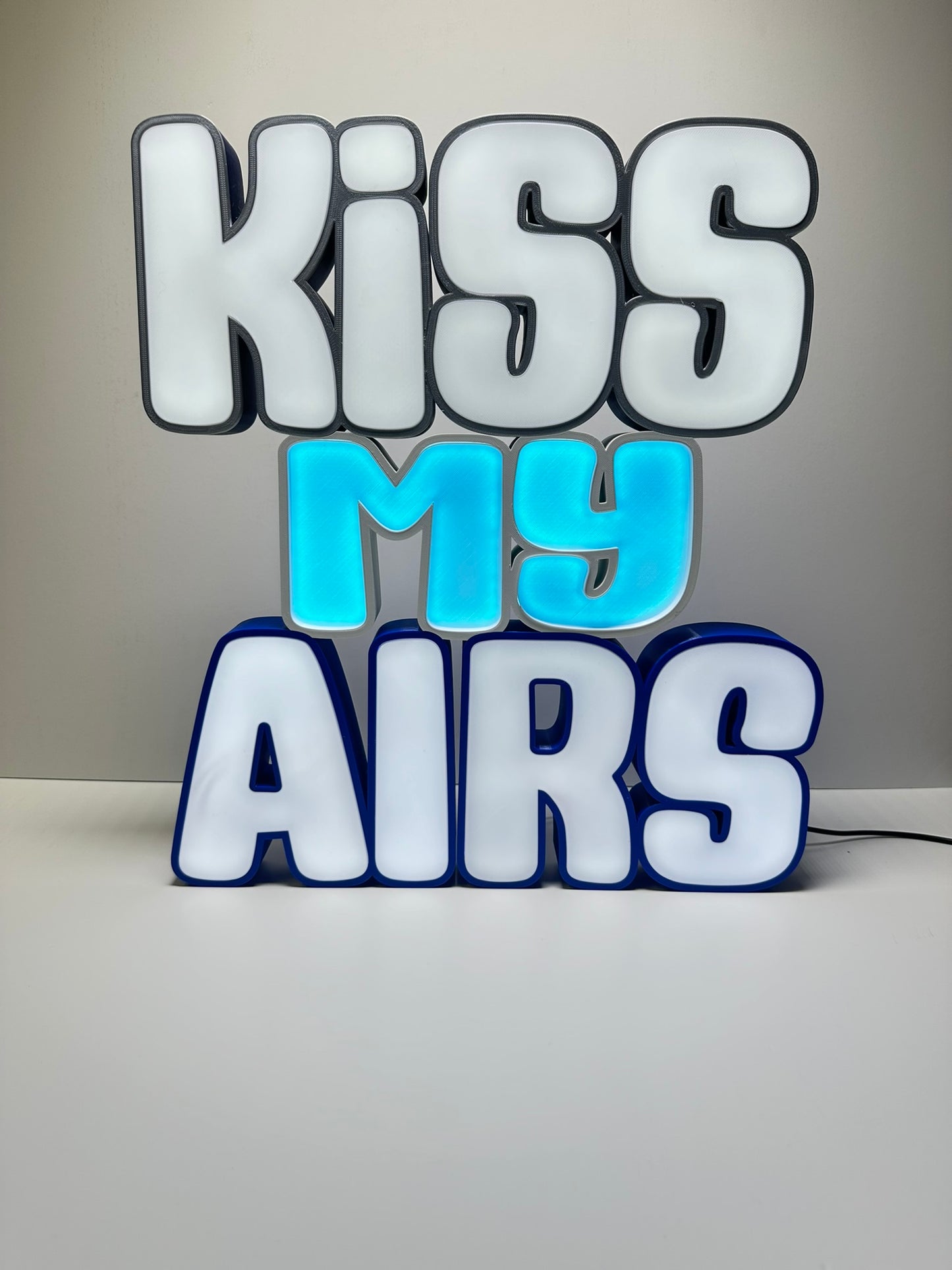 Front view of the Kiss My Airs LED Channel Letter Sign, featuring bold white letters with black outlines and a light blue 'MY' in the center, illuminated against a white background.