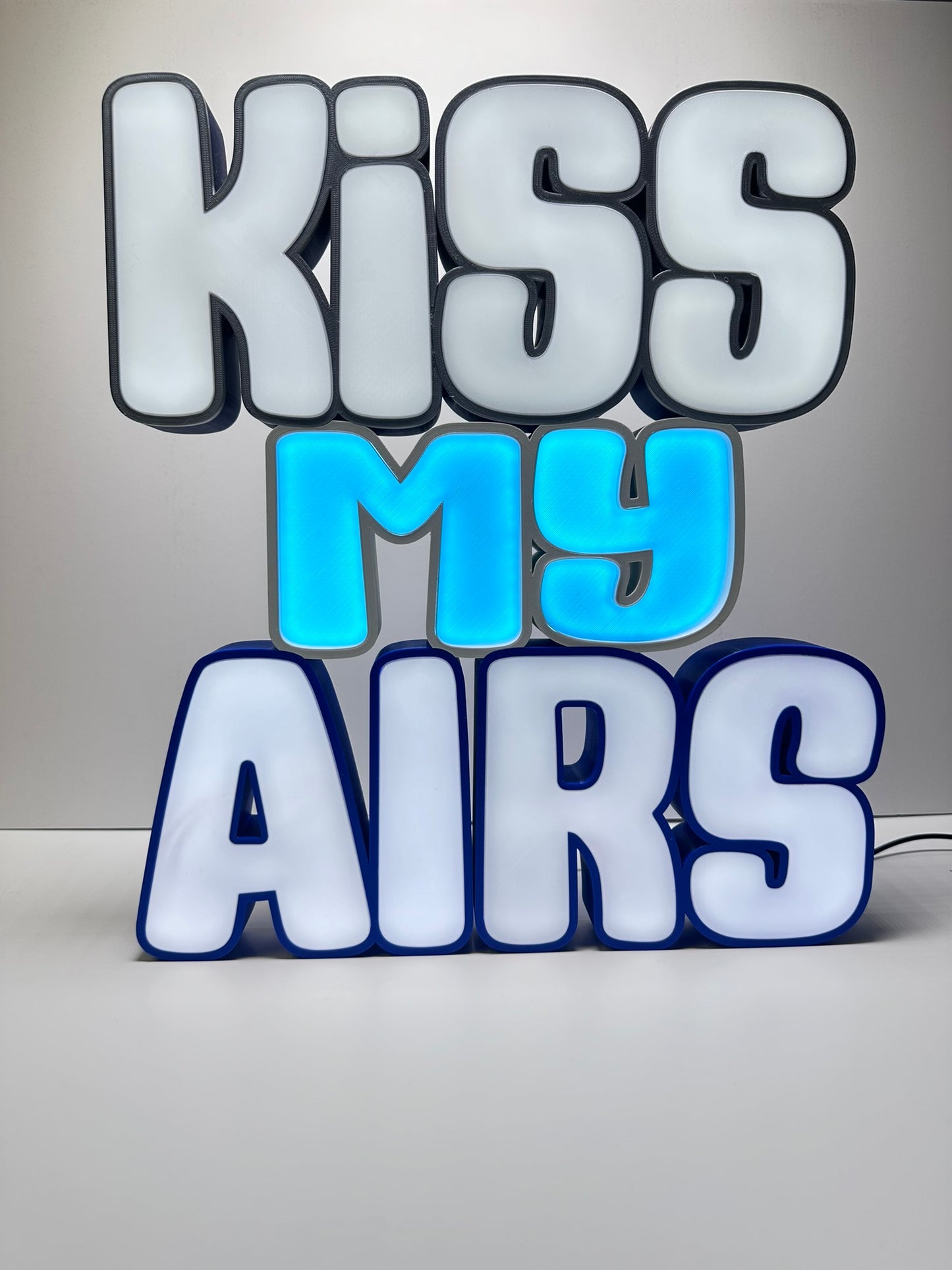 Front view of the Kiss My Airs LED Channel Letter Sign, featuring bold white letters with black outlines and a light blue 'MY' in the center, illuminated against a white background, perfect for home or office decor.