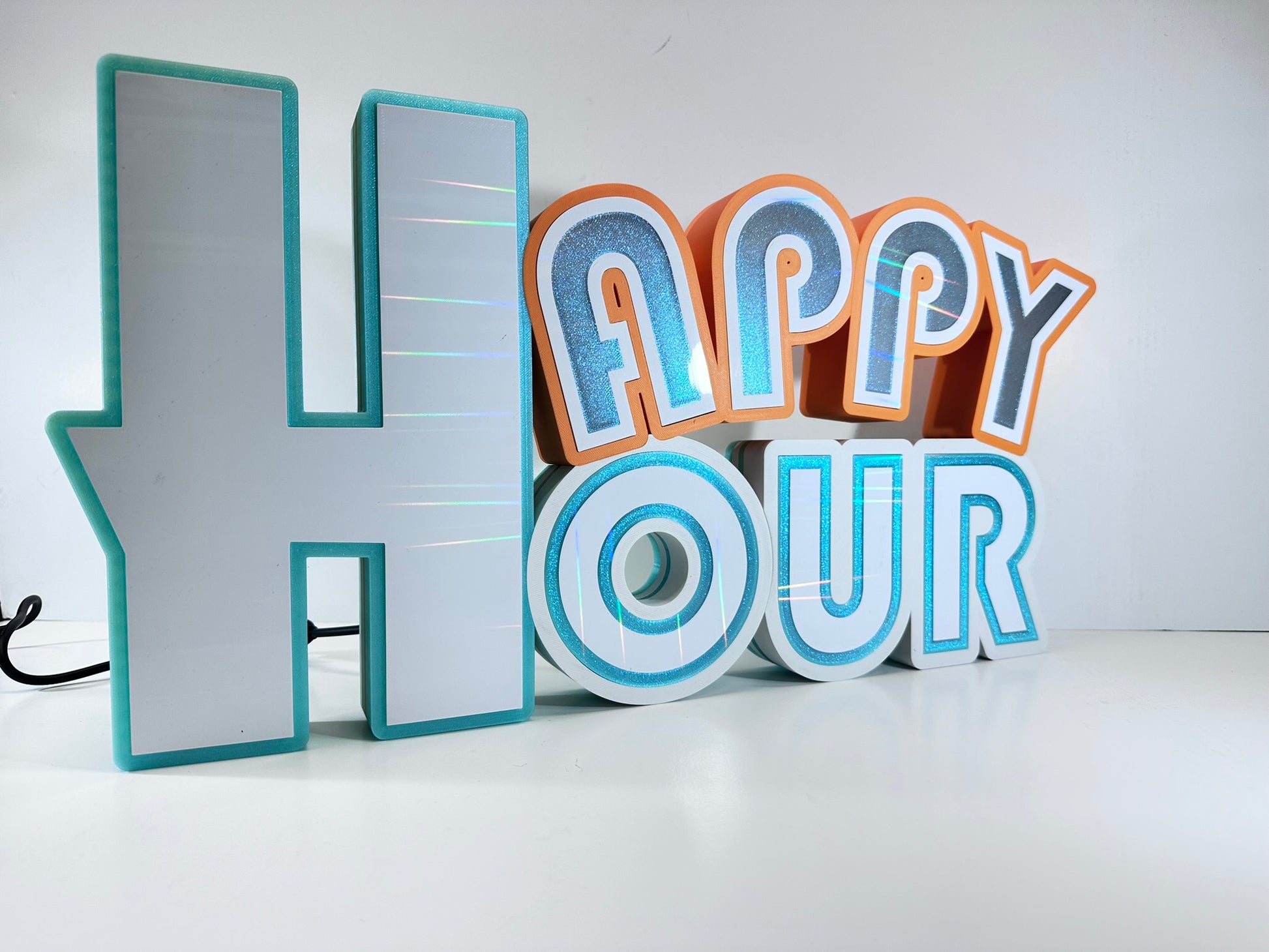 Side angle of a 'Happy Hour' 3D printed sign showing the depth and texture of the letters with turquoise and orange details on a white surface