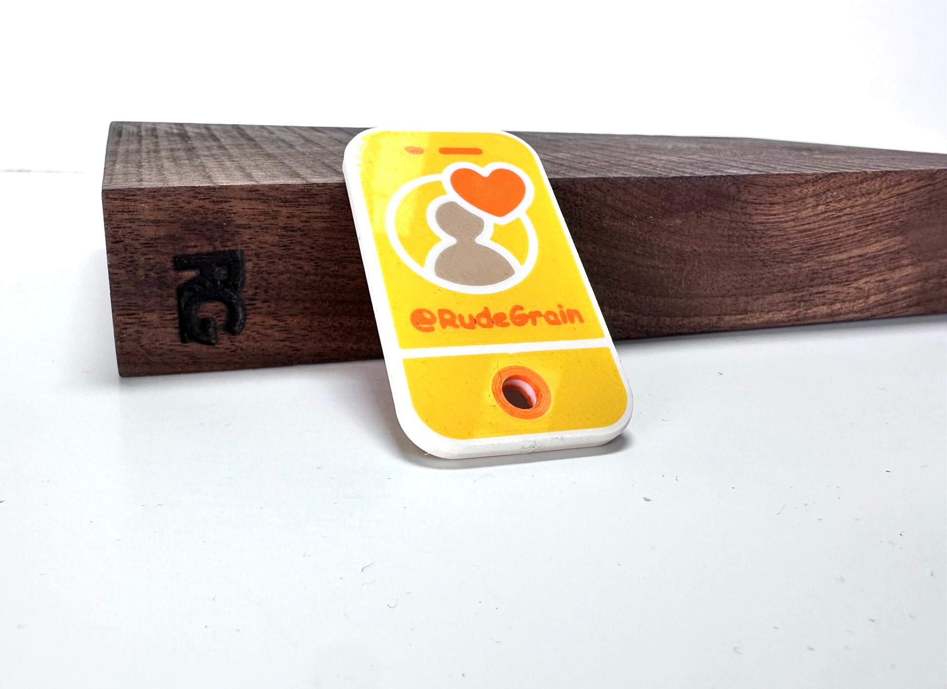 Yellow NFC/QR tag featuring a heart icon and the @RudeGrain branding, displayed on a wooden stand with engraved RG initials