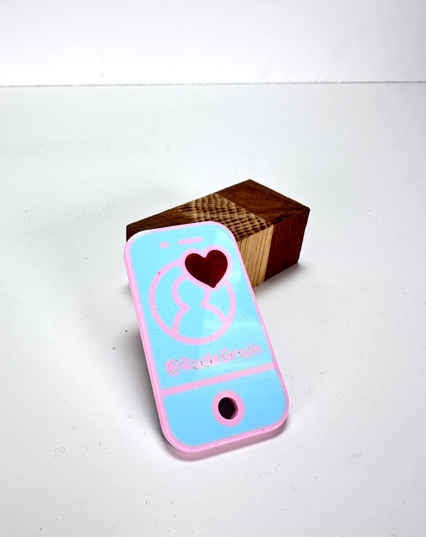 Pink and blue NFC/QR tag with a heart icon and @RudeGrain branding, resting on a natural wooden block.